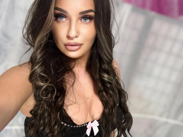 SweetSerena-hot with long wavy brown hair, blue eyes, and pink lipstick, wearing a black top with a pink bow, showing her cleavage. She has a cute nose and full lips.