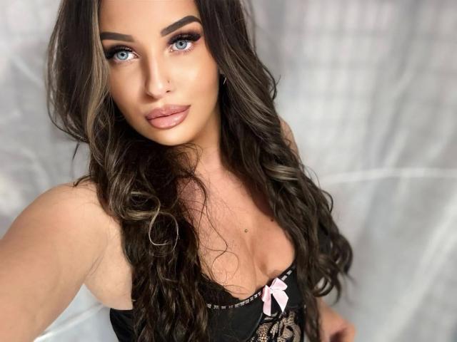 SweetSerena-hot with long, wavy brown hair, blue eyes, and pink lipstick, wearing a black lace top with a pink bow, looking at the camera with a confident expression.