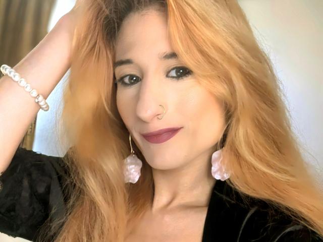 SeductiveBonita with blonde hair, nose ring, purple lipstick, black dress, pink earrings, white bracelet, smiling, hand in hair, soft light, gentle expression.