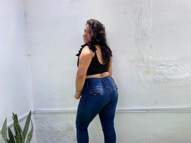 Ana Jimenez, con i capelli neri ricci, una maglietta nera con volant e jeans blu attillati, mostra il sedere e sorride alla telecamera. In basso a sinistra, una pianta verde..