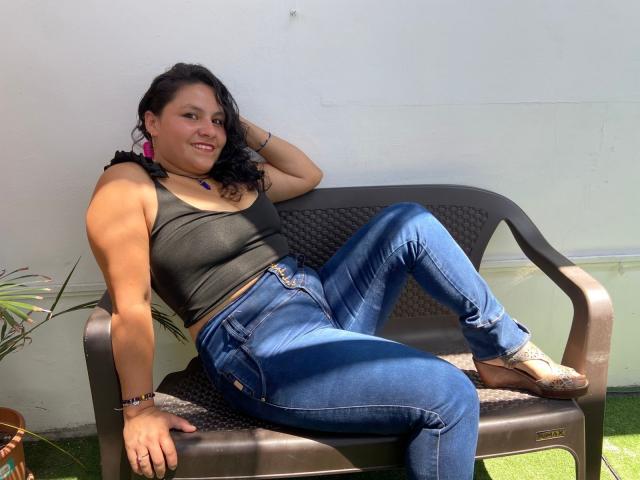 Ana Jiménez, con cabello oscuro, aretes rosas, un top corto negro, jeans azules y botas color beige, está recostada en un banco marrón, sonriendo, con un brazo detrás de la cabeza y las piernas cruzadas..