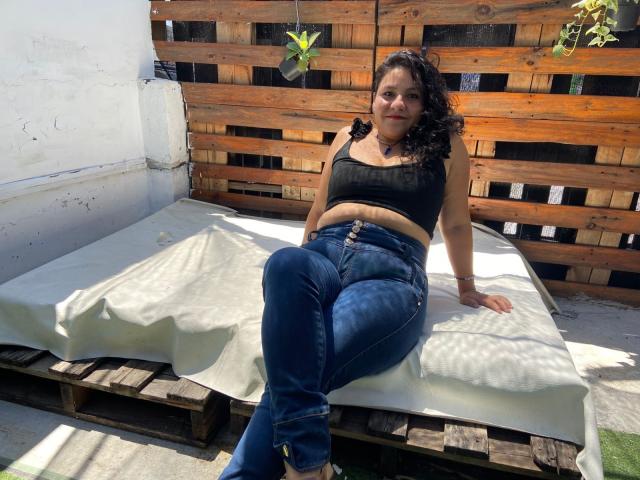 AnaJimenez, com cabelos pretos encaracolados, seios médios usando uma cropped preta e jeans azuis justos, sentada em uma palete de madeira com um colchão branco, ao ar livre..