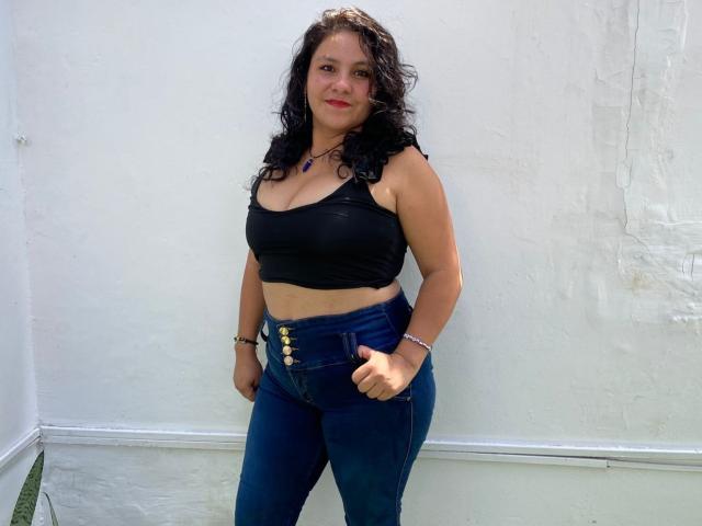 Ana Jimenez mit lockigem, schwarzem Haar, einem schwarzen Tanktop, das ihre Brüste zeigt, und hoch taillierten, blauen Jeans mit goldenen Knöpfen, steht vor einer weißen Wand und lächelt leicht..