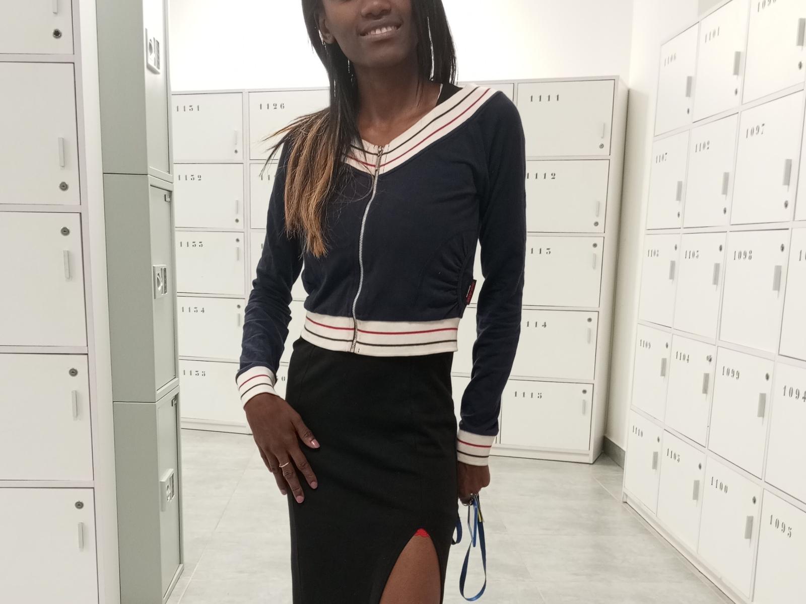 Nanou69s with dark skin, long black hair, wearing a navy blue long-sleeve top with red and white stripes, black skirt with red slit, standing in locker room.