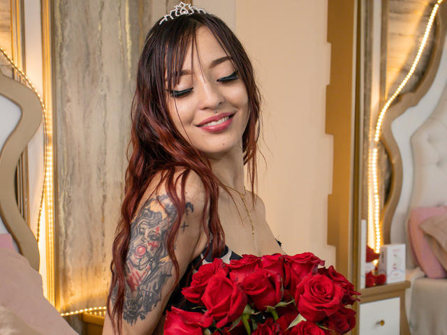 CatalleyaDubois with red roses, smiling, wavy brown hair, tattooed arm, silver tiara, holding bouquet, bare shoulders, pink lipstick, soft lighting, bedroom setting.