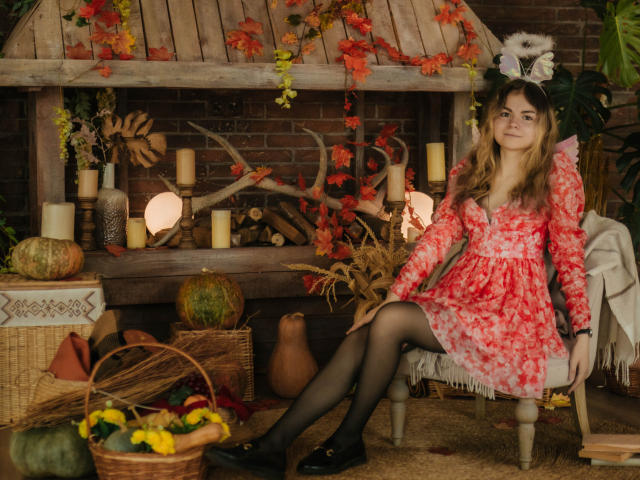 TinaRedly with autumn vibes, wearing a red floral dress, black stockings, and black flats, sitting on a wooden chair, surrounded by pumpkins, candles, and fall decorations.