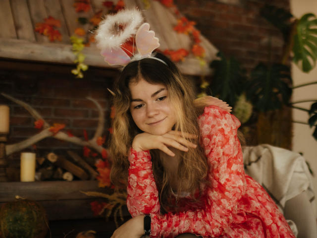 TinaRedly with long blonde hair, wearing a pink floral dress and fluffy white angel wings, smiling softly, hands under chin, sitting in a cozy, autumn-themed room.