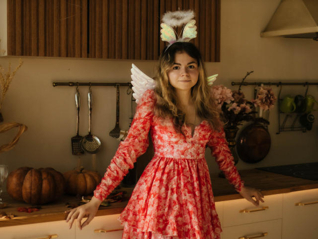 TinaRedly, wearing a red floral dress with white angel wings and fluffy halo, stands in a kitchen, her hands on the counter, showing a cute, sexy, angelic look.