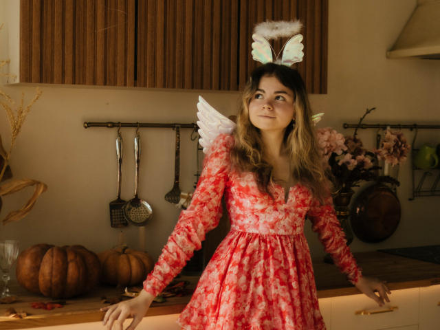 TinaRedly with long brown hair, wearing a red floral dress and white fairy wings, has small tits and is leaning on a kitchen counter with wooden cabinets.