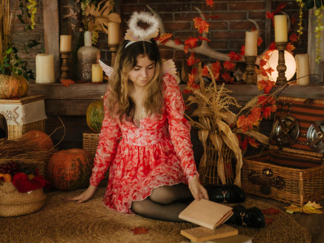 TinaRedly mit welligem, blondem Haar, einem roten Kleid mit Blumenmuster, weißen Engelsflügeln und einem Heiligenschein, sitzend auf Heu, schwarzen Schuhen, einem Buch und Kürbissen, umgeben von Kerzen und Herbstlaub..