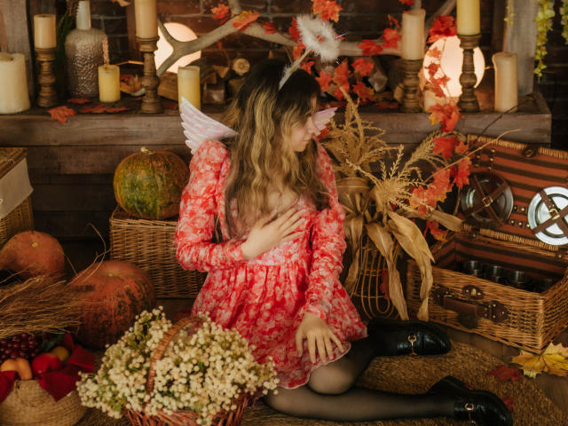 TinaRedly mit welligem, blondem Haar, einem roten Kleid mit Blumenmuster, weißen Flügeln und einem flauschigen Kopfschmuck, kniet und berührt ihre Brüste, umgeben von Kerzen, Herbstlaub und Früchten, und trägt schwarze Stiefel..