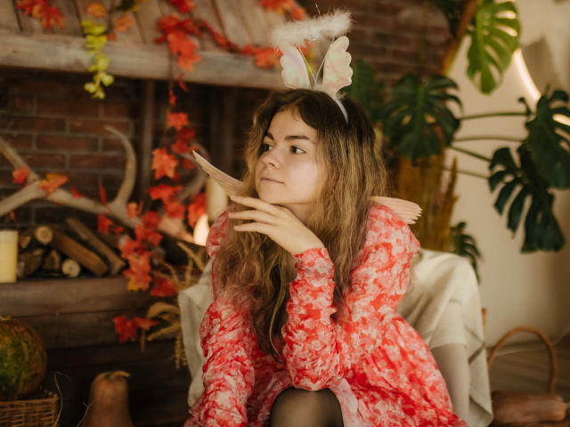 TinaRedly with wavy brown hair, wearing a red floral dress, white bunny ears, and a fluffy scarf, sitting, looking thoughtful, hands on chin, warm indoor setting.