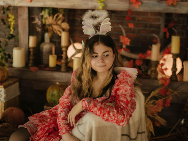 TinaRedly, com cabelos castanhos ondulados, vestindo um vestido floral vermelho e orelhas de coelho brancas, sentada em uma cadeira de madeira, sorrindo, cercada por velas e folhas de outono..