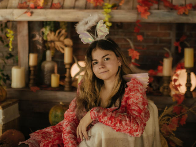 TinaRedly with long brown hair, wearing a red floral dress and white fluffy headband, sitting in a cozy room with candles and autumn leaves, smiling gently at the camera.