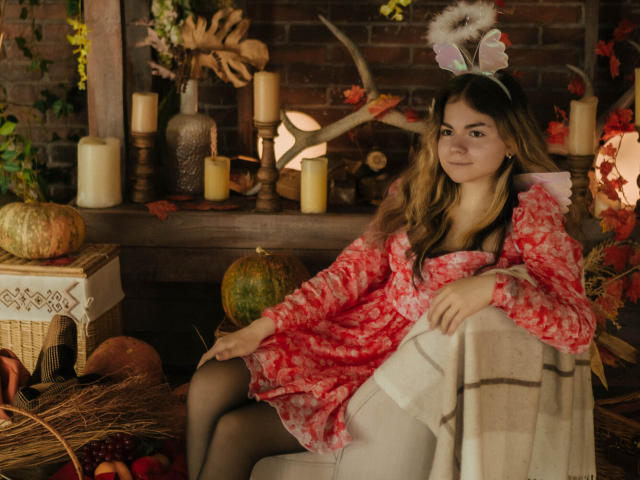 TinaRedly, with long brown hair, wears a red floral dress, black tights, and bunny ears, sitting on a chair, surrounded by candles, pumpkins, and a plaid blanket.