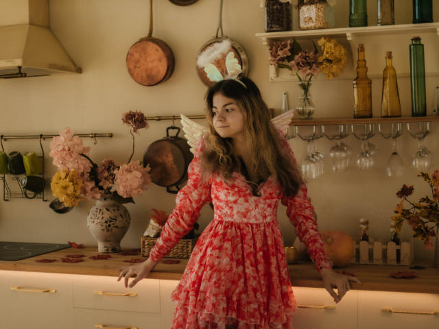 TinaRedly vestindo um vestido floral vermelho com asas brancas, cabelo castanho e orelhas de coelho, apoiada em uma bancada de cozinha com flores rosa e amarelas, panelas de cobre e copos pendurados..
