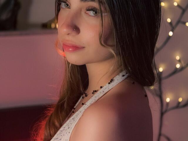 AntonellaFerrara has long brown hair, a nose piercing, and wears a white halter top with black bead accents. Her red lips and green eyes are softly lit.