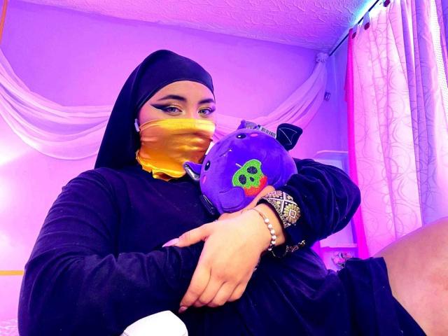 AishaGhulam in a black hijab and yellow mask, holding a purple stuffed animal with a green "B" on it, wearing a black dress and bejeweled bracelet.