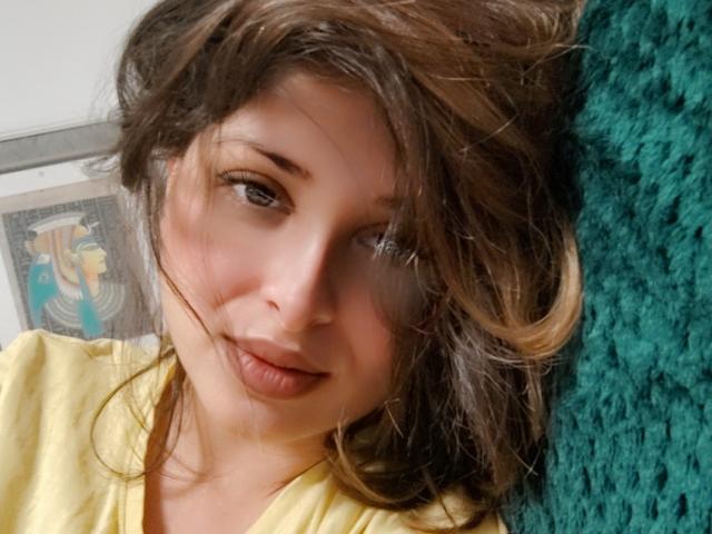 ScarletOra with messy brown hair, pouty lips, and a cute smile, wearing a yellow shirt, lying against a green textured pillow. Her tits are not visible.