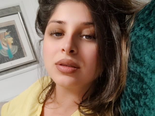ScarletOra met bruin haar, groene ogen en roze lippen, die een gele blouse draagt en lichtelijk opzij kijkt, op een groen, gestructureerd oppervlak. Haar gezicht is gecentreerd in het beeld..