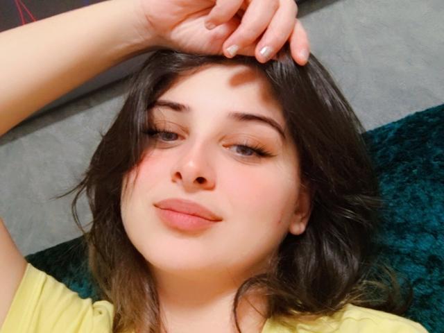 ScarletOra met donkerbruin haar, lichte huid en volle lippen, liggend, met haar hand op haar hoofd, gekleed in een geel shirt, met een lichte glimlach, met een groene en grijze achtergrond..