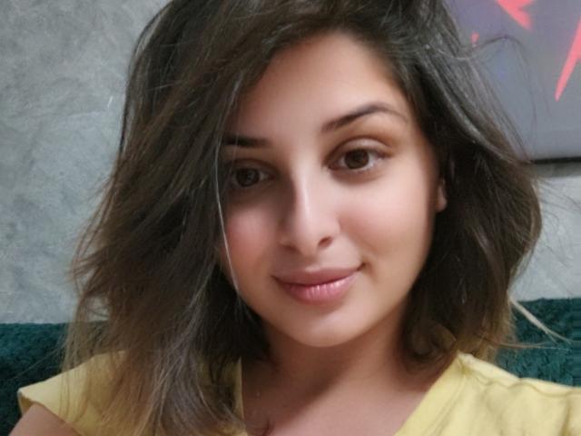ScarletOra with wavy brown hair, light skin, and a subtle smile, wearing a yellow shirt, has cute brown eyes and a small nose, looking slightly to the right.