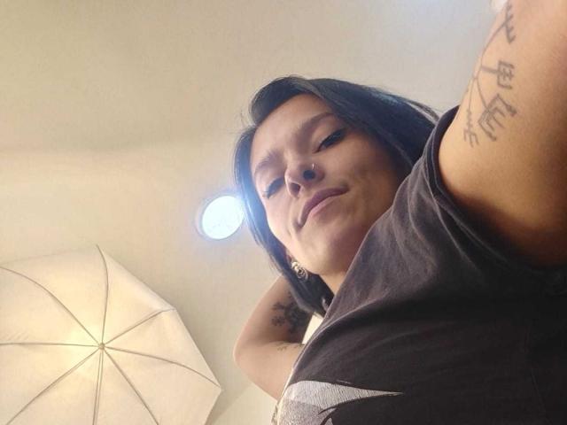 Amelie Blake, con cabello negro, sonriendo hacia la cámara, vistiendo una camisa gris, mostrando tatuajes en su brazo, bajo una luz de techo y un paraguas..