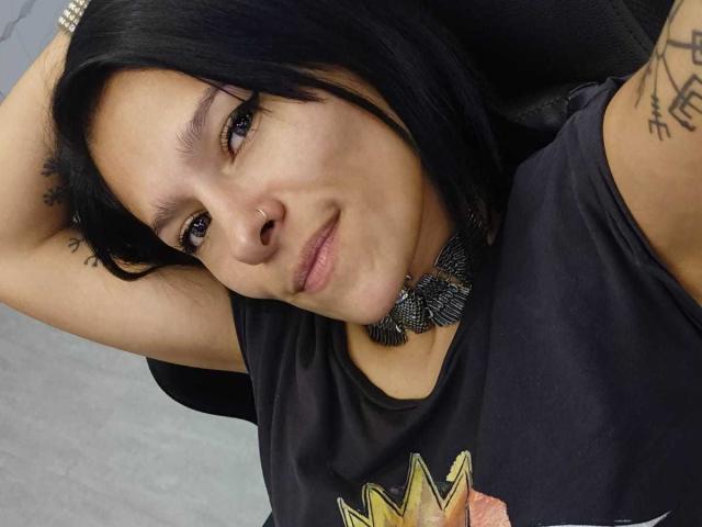 AmelieBlake has black hair, a nose ring, and a tattoo on her arm. She's wearing a black shirt and a chunky necklace, with arms raised, looking at the camera.