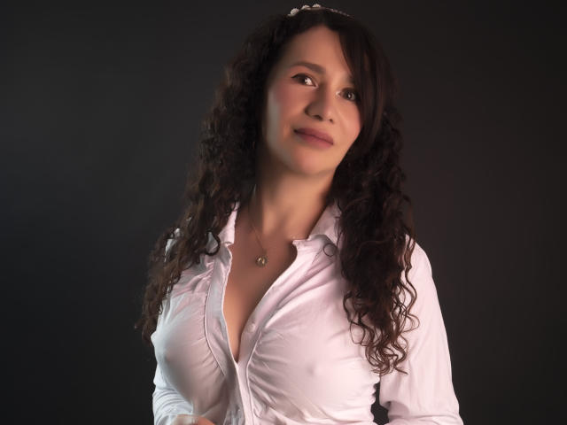 LizzieAnderson with curly dark hair, wearing a slightly unbuttoned white shirt, showing her tits, looking up with a subtle smile, dark background, soft lighting.