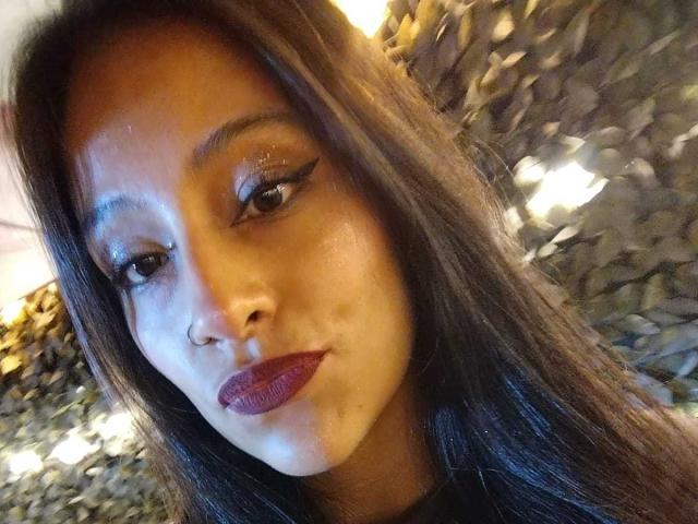 NoremCute has dark brown hair, shiny blue eyeshadow, and deep red lipstick, with a slight pout, looking into the camera. She's wearing a nose ring.