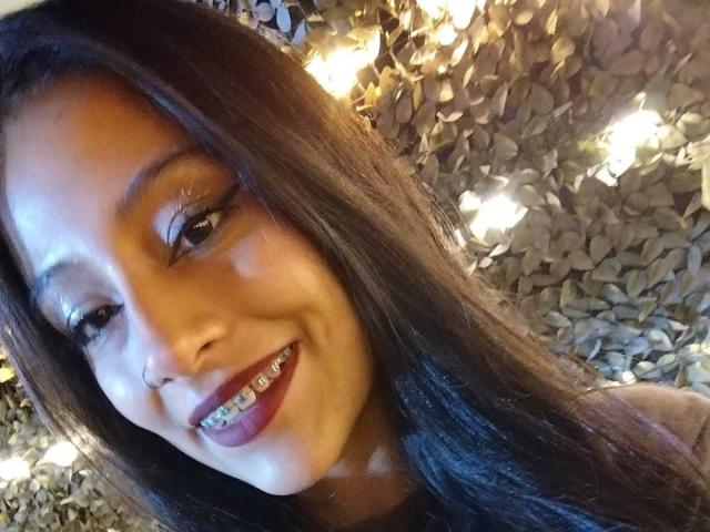 NoremCute has brown skin, long black hair, and a smile showing metal braces. She's looking at the camera with warm, glowing lights in the background.