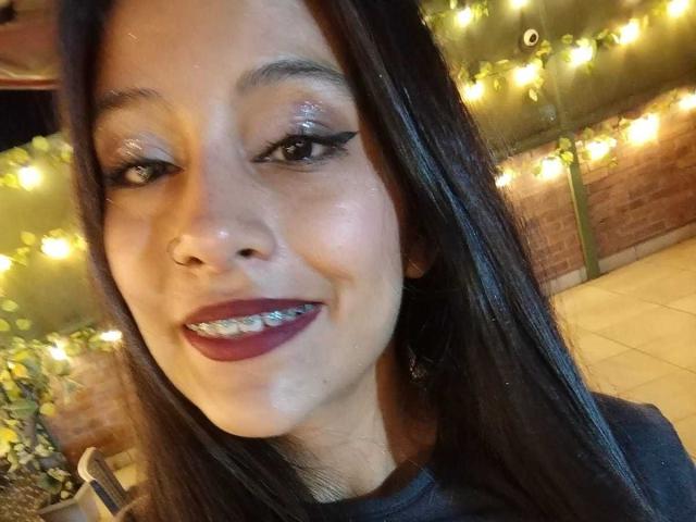 NoremCute has shiny brown eyes, long black hair, and is smiling with dark red lipstick, showing metallic teeth. She's wearing a nose ring and a black top.
