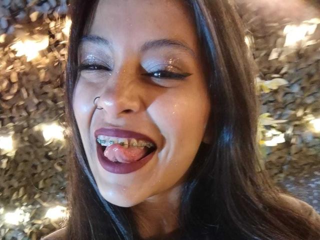 NoremCute with dark hair, nose ring, and glittery eyeshadow, tongue out, showing metal braces, and wearing burgundy lipstick, smiling with a slightly shiny face.