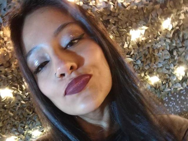 NoremCute with shiny blue eyeshadow, dark purple lipstick, and long brown hair, pouting with a slight smile, nose pierced, standing against a blurred, leafy background.
