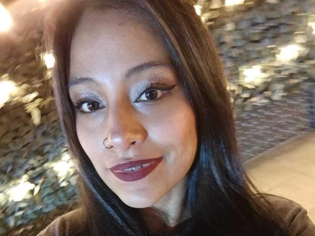 NoremCute has dark brown hair, a nose ring, and dark purple lipstick. She's smiling with sparkly eye makeup and looking at the camera. Her tits aren't visible.