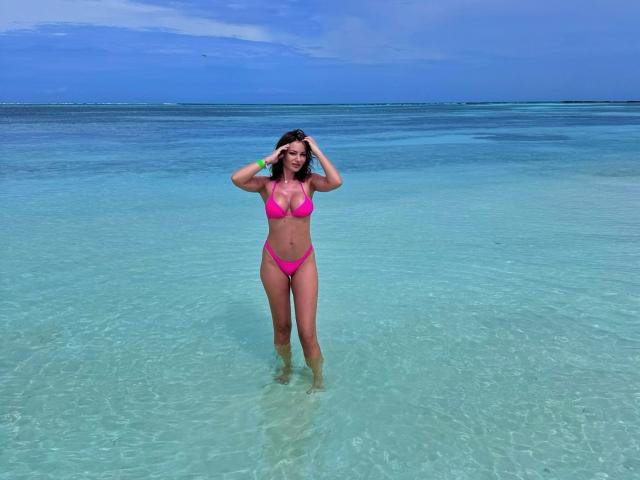 AliceMarr with pink bikini, standing in clear blue water, hands in her brown hair, showing her tits and ass, smiling, sunlight on her tan skin, ocean horizon.