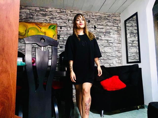 Sara Duncan está de pie en una habitación, vestida con una camiseta negra que deja ver sus piernas tatuadas. Tiene el cabello castaño liso, lápiz labial rojo y una expresión segura. Está cerca de una silla negra..