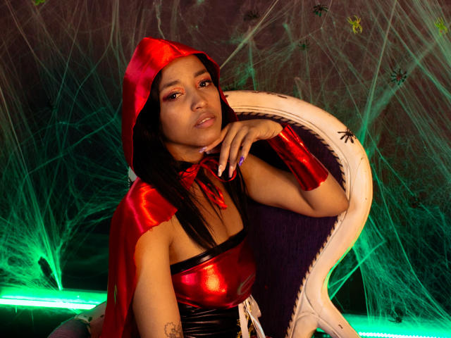 ShanthalBlizz with brown skin, red hood, and shiny red top, sitting in a white chair, wearing a red cape and armlet, green light, and spider web background.
