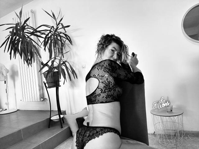 AngeelCouple with curly hair, wearing black lace lingerie, sits on steps, showing her ass, holding a pen to her mouth, looking back, in a black-and-white photo.