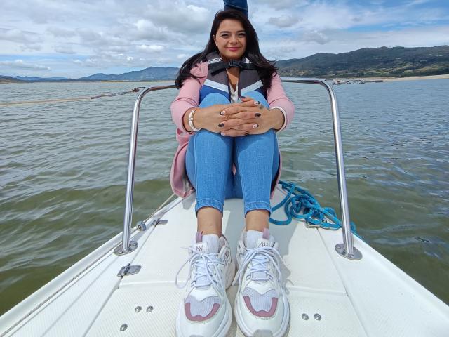 katherinLatin is sitting on a boat with her knees up, wearing a pink jacket, blue jeans, and white sneakers, hands on her knees, looking at the camera.