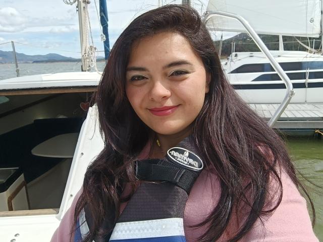 Katherin Latin heeft lang, donkerbruin haar, roze lippenstift en een schattige glimlach. Ze draagt een zwarte reddingsvest over een roze shirt en staat op een boot..