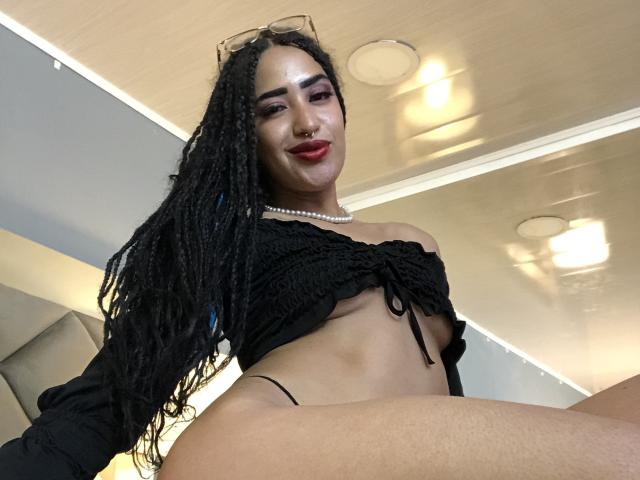MichellDiaz with long black braids, black off-shoulder crop top, pearl necklace, red lipstick, and black thong, sitting with legs apart, showing her smooth, tan, bare ass.