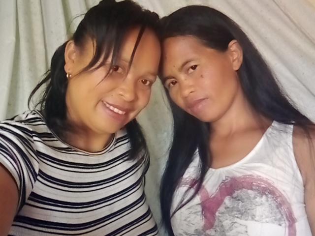 Two sexy Black women with long black hair, smiling and posing together. One wears a striped shirt, the other a white tank top with a pink design.