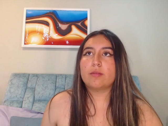 SalomettX with long brown hair, looking up, has a chubby face, small tits, and is shirtless, sitting in front of a blue-patterned headboard and a colorful abstract painting.