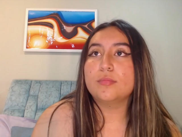 SalomettX with long brown hair, brown eyes, and light skin, looking up with a neutral expression, showing small zits on her nose and upper lip. Her boobs are not visible.