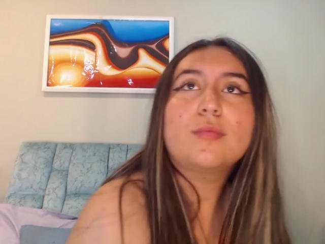 SalomettX has long brown hair, full lips, and a small nose. She's looking up with a slightly pouty expression. Her bare shoulders are visible. A colorful abstract painting is behind her.