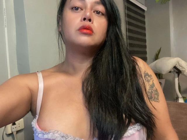 YENDYSFOX01-hot with long black hair, red lips, light skin, pink lace bra showing small tits, tattoo on right shoulder, looking at camera. Indoor setting.