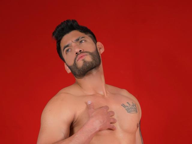 Shirtless BadHotGuys69 with a dark beard, short black hair, and a tattoo on his chest, giving a thumbs-up, against a solid red background. His chest is hairy.