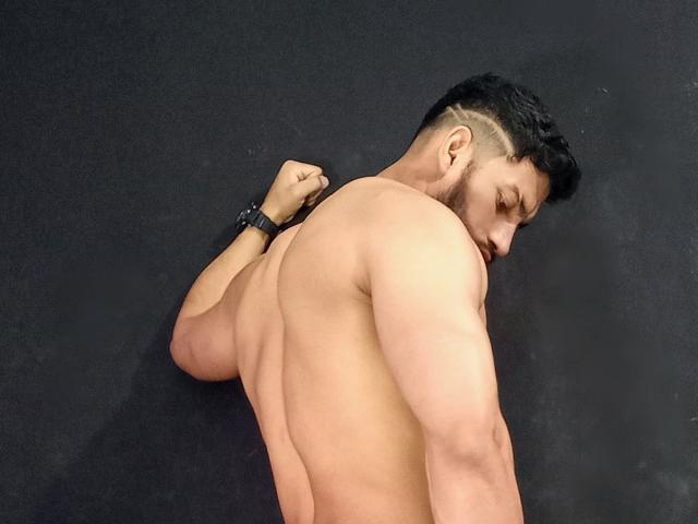 BadHotGuys69, shirtless, muscular, tan skin, black hair, undercut, wristband, butt raised, looking back, right arm bent, black background, standing, serious expression, strong back, defined muscles.