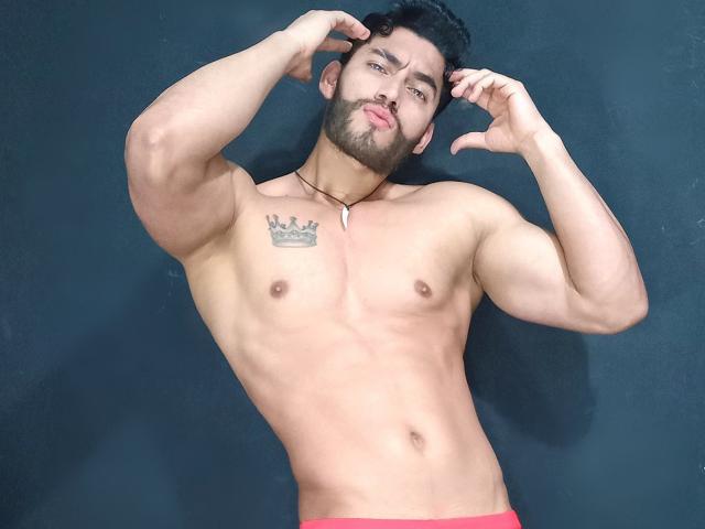 Muscular BadHotGuys69 with black curly hair and beard, flexing arms, chest tattoo, necklace, and red briefs, standing against dark background, showing toned abs and pectorals.